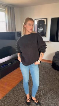 Jauna sieviete stāv viesistabā ar rokām kabatās, valkā melnu džemperi, gaiši zilas džinsas, melnas kurpes un viedpulksteni uz kreisās rokas.