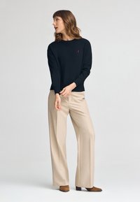 Suéter negro con cuello redondo, puños acanalados y logotipo rojo. Combinado con pantalones beige de pierna ancha y botas de tobillo con tacón marrones.