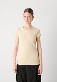 Kremfarget ribbestrikket t-skjorte med kort erme og rund hals, tettsittende design, og subtil broderidetalj på brystet, båret sammen med svarte bukser.
