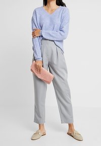 Jasnoniebieski sweter prążkowany z dekoltem w kształcie litery V, zestawiony z jasnoszarymi spodniami w kant. Trzyma różową torebkę typu kopertówka, beżowe buty wsuwane z akcentami.