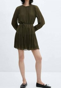 Oliivinvihreä laskostettu mekko, jossa on pitkät hihat, istutettu vyötärönauha ja kerroksellinen helma, yhdistetty mustiin mataliin kenkiin.