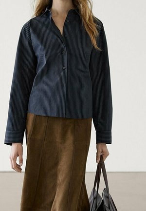 Femme portant une chemise bleu marine à fines rayures et une jupe en daim marron, tenant un sac cabas en cuir noir dans sa main droite.