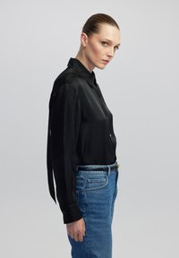 Schwarze Seidenbluse mit langen Ärmeln, Knopfleiste und klassischem Kragen, kombiniert mit hellblauen Jeans. Glatte Textur und taillierter Schnitt.