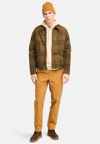 Chaqueta de cuadros marrón, sudadera beige debajo, pantalones color camel, botas marrones y un gorro mostaza. Presenta dos bolsillos frontales y detalles en cremallera.