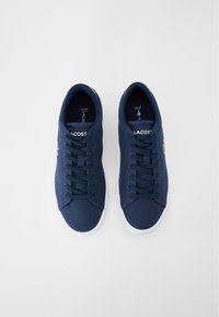 Zapatillas de lona de color marino con puntera redonda, suela de goma blanca y cordones en contraste; presenta el logo de Lacoste en el costado y una superficie texturizada.