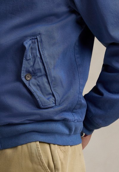 Primer plano de un bolsillo con solapa abotonada de una chaqueta azul, dobladillo acanalado y puño de manga, llevado sobre pantalones beige.