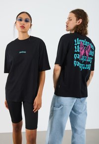 Black oversized t-shirt featuring small front text and large colorful butterfly graphic with text on the back; worn with denim and black shorts.