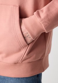 Rosa Sweatshirt mit Kängurutasche, gerippten Bündchen und Saum. Weiche Textur mit sichtbaren Nähdetails rund um die Tasche.