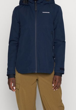 Persoon draagt een marineblauwe Didriksons capuchonjack met ritssluiting en lichtbruine cargobroek, staand tegen een effen achtergrond.