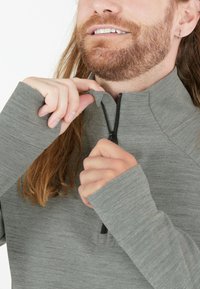 Harmaa pitkähihaainen neule, jossa on neljännesvetoketju, sileä kangas, peukalonreikä hihansuissa ja teksturoitu pinta. Musta vetoketj yksityiskohtana.