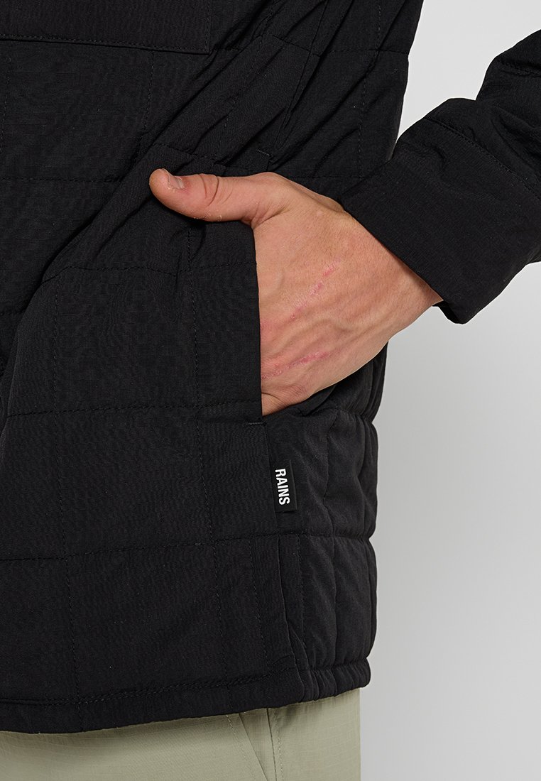 Hand med synliga ärr inskjuten i framfickan på en svart quiltad RAINS-jacka.