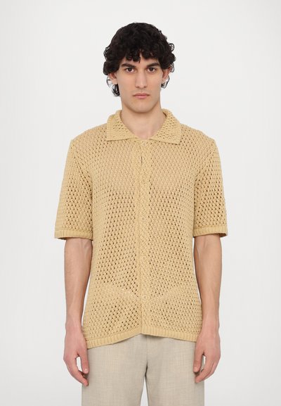 Chemise à manches courtes en tricot beige avec un col uni, fermeture à boutons et un motif en maille ouverte. Portée avec un pantalon de couleur neutre.