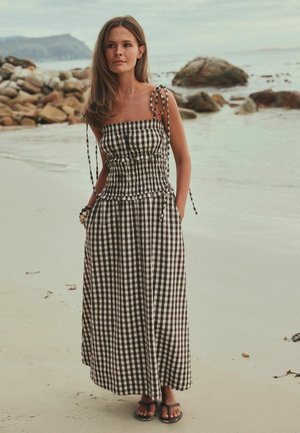 Ung kvinde står på en sandstrand iført en sort-hvid ternet kjole med skulderbånd og sandaler, med klipper og vand i baggrunden.