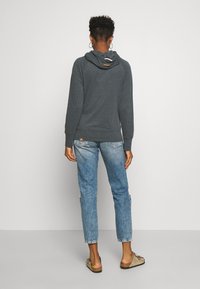 Grijze hoodie met een afgeronde zoom en trekkoordkap, gecombineerd met lichtblauwe versleten jeans en brown instapschoenen.