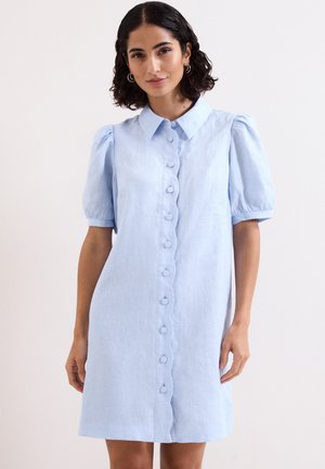 Vrouw met kort krullend haar die een lichtblauwe jurk met korte mouwen en knoppen aan de voorkant draagt, staand tegen een simpele achtergrond.