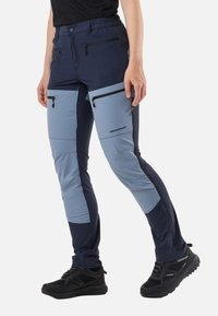 Person wearing two-tone blue hiking pants with multiple zip pockets and black waterproof hiking shoes, standing with one leg forward.