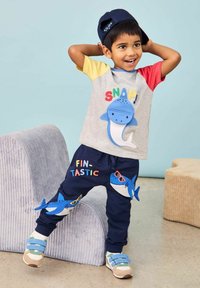 Harmaa paita, jossa on värikäs "SNAP!"-teksti ja sininen hain kuvio, yhdistettynä tummiin housuihin, joissa on hai-aplikointeja ja värikäs "FIN-TASTIC"-teksti.