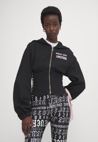 Čierna skrátená mikina s kapucňou a zipsom, s nadrozmernými rukávmi a vyšívaným nápisom "VERSACE JEANS COUTURE" v ružovej farbe. V kombinácii s čiernymi legínami s bielou typografiou.