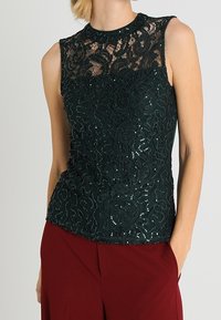 Top sin mangas de color verde oscuro con encaje floral y lentejuelas, cuello alto y forma ajustada, combinado con pantalones burdeos.