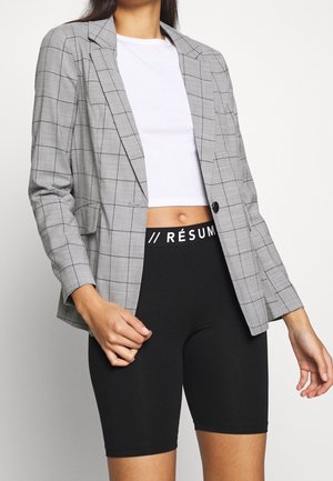Nainen, jolla on yllään harmaa ruudullinen bleiseri, valkoinen lyhyt t-paita ja mustat korkeavyötäröiset pyöräilyshortsit, joissa on "RÉSUM" vyötärötekstinä.