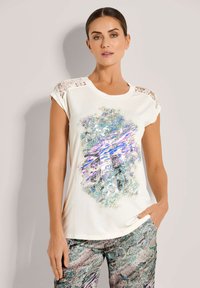 Vrouw in een crème T-shirt met korte mouwen en kanten schouders, met een abstract blauw en paars print, gecombineerd met een patroonbroek, staande voor een effen muur.