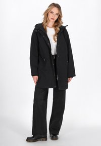 Parka nera impermeabile con cappuccio, dotata di bottoni a pressione e zip frontale, abbinata a pantaloni neri a gamba larga e scarpe nere chunky.