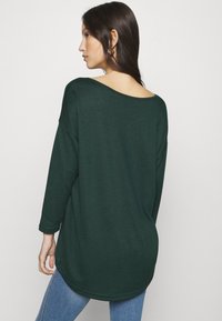 Mørkegrøn oversized sweater med bred udskæring og trekvartærmer, prydet med en rundet kant og blødt tekstureret stof.