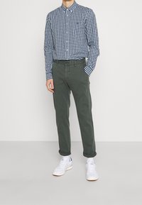 Camicia a quadri blu e bianca con bottoni abbinata a pantaloni chino verde oliva e sneakers bianche. Maniche dei pantaloni arrotolate, vestibilità casual.