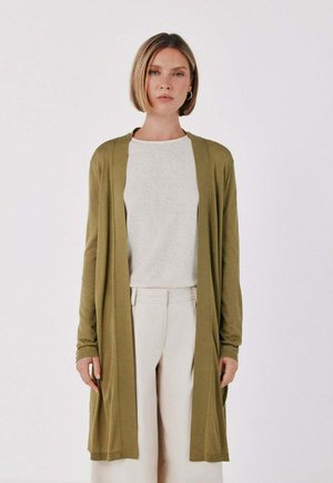 Donna con capelli corti biondi che indossa un cardigan aperto verde oliva, una maglietta grigio chiaro e pantaloni beige chiaro, in piedi davanti a uno sfondo semplice.