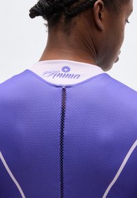 Κοντινό πλάνο του πάνω μέρους της πλάτης ενός ατόμου που φοράει μωβ αθλητική μπλούζα με υφή και λευκό γιακά με την ένδειξη "Anima" και σκούρα πλεξούδα μαλλιών δεμένα ψηλά.