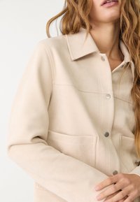 Jasnobeżowa kurtka z miękkiego materiału, z kołnierzem, zatrzaskami i dwoma przednimi kieszeniami z minimalnymi przeszyciami.