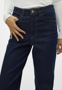 Mörkblå denim högmidjade jeans med orange stygn, en framficka och en silverknappsstängning vid midjan.