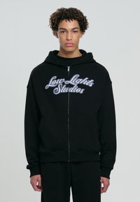 Giovane con capelli ricci che indossa un hoodie nero con zip con la scritta "Low Lights Studios" sul davanti, su uno sfondo semplice.