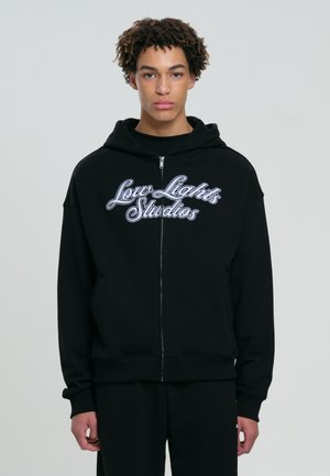 Noor curly juuksed noor inimene, kellel on seljas must tõmmatav kapuuts, mille ees on kiri "Low Lights Studios", taustal on ühtlane taust.