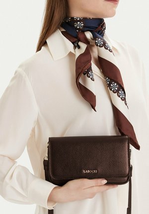 Mujer con blusa blanca y pañuelo con estampado sostiene un bolso de mano de cuero marrón oscuro de Lasocki a la altura de la cintura.