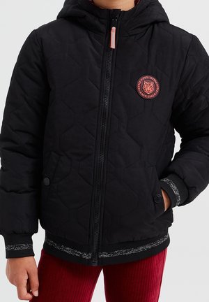 Dziecko ubrane w czarną, pikowaną kurtkę z naszywką z logo tygrysa oraz czerwone spodnie, z rękami w kieszeniach, na jednolitym tle.