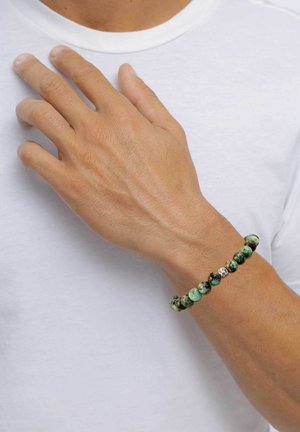 Beaded bracelet featuring various shades of green and black stones, interspersed with a silver accent, worn on a hand against a white shirt.