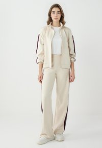 Beige Jogginganzug mit einer Reißverschluss-Bomberjacke und weitem Beinkleid, das tief weinrote Seitenstreifen aufweist. Weicher Stoff und lockere Passform.