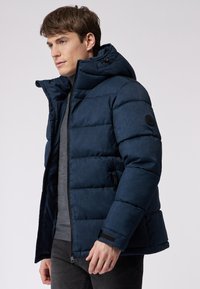 Mørkeblå puffervintersjakke med struktureret overflade, hætte, lynlås og sidelommer. Har velcro-detaljer på ærmerne.