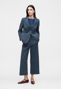 Femme debout les mains dans les poches portant un costume bleu marine à fines rayures, un pantalon large et court, et des ballerines marron Mary Jane.