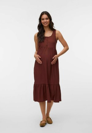 Tehotná žena stojí a usmieva sa, má na sebe hnedé midi šaty bez rukávov a hnedé rovné topánky, ruky jemne spočívajú na bruchu.