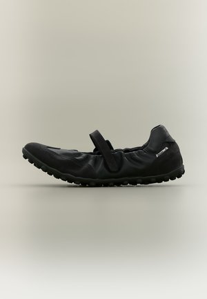 Chaussure noire flexible style Mary Jane avec une bride à scratch et une semelle texturée, vue de côté sur un fond neutre.