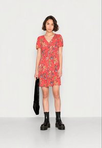 Vestido floral vermelho com mangas curtas em balão, decote em V e cintura ajustada. Botas pretas largas a fazer o tornozelo e uma mala preta segurada na mão esquerda.