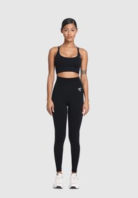 Žena stojí čelom dopredu, oblečená v čiernom športovom podprsenku, vysokých čiernych legínach so štítkom, bielych teniskách a viditeľnými tetovaniami na ramenách na šedom pozadí.