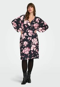 Žena s dugom kosom koja nosi crnu haljinu s ružičastim cvjetnim uzorkom, crne tajice i crne kratke čizme, pozira s rukama na bokovima.