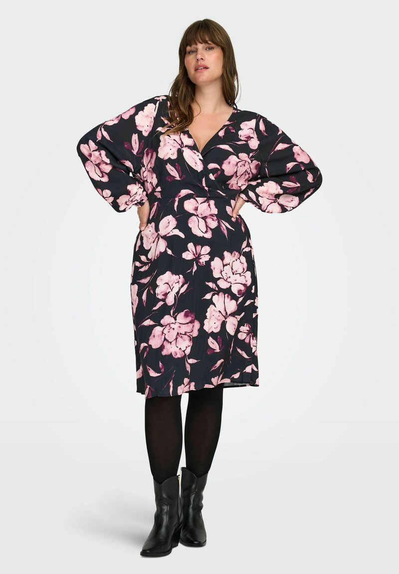 Žena s dugom kosom koja nosi crnu haljinu s ružičastim cvjetnim uzorkom, crne tajice i crne kratke čizme, pozira s rukama na bokovima.