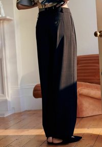 Personne portant un pantalon taille haute à jambes larges avec un motif à carreaux foncé, debout à l'intérieur sur un sol en bois près d'un canapé couleur rouille.