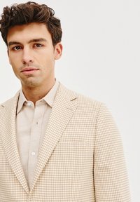 Beżowa marynarka w krate houndstooth o dopasowanym kroju i wyraźnych klapach, w połączeniu z jasnobeżową koszulą z guzikami, posiadającą subtelną fakturę.