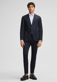 Marineblauw op maat gemaakt pak met een enkelvoudig jasje, twee voorzakken en slim-fit broek, gecombineerd met een lichte gedessineerde blouse en zwarte instappers.