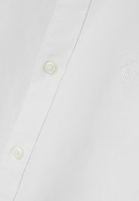 Camicia bianca con bottoni a pressione, caratterizzata da una trama a tessuto semplice, con un bottone tondo in plastica e un logo ricamato sul lato sinistro.